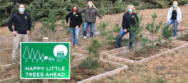 a small group of masked women and men stand with planted trees and a Happy Little Trees Ahead green sign at Higgins Lake State Park
