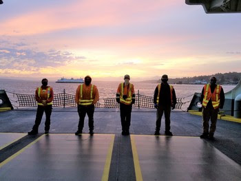 Photo of employees lined up on car deck of ferry with sunrise and another ferry in the background