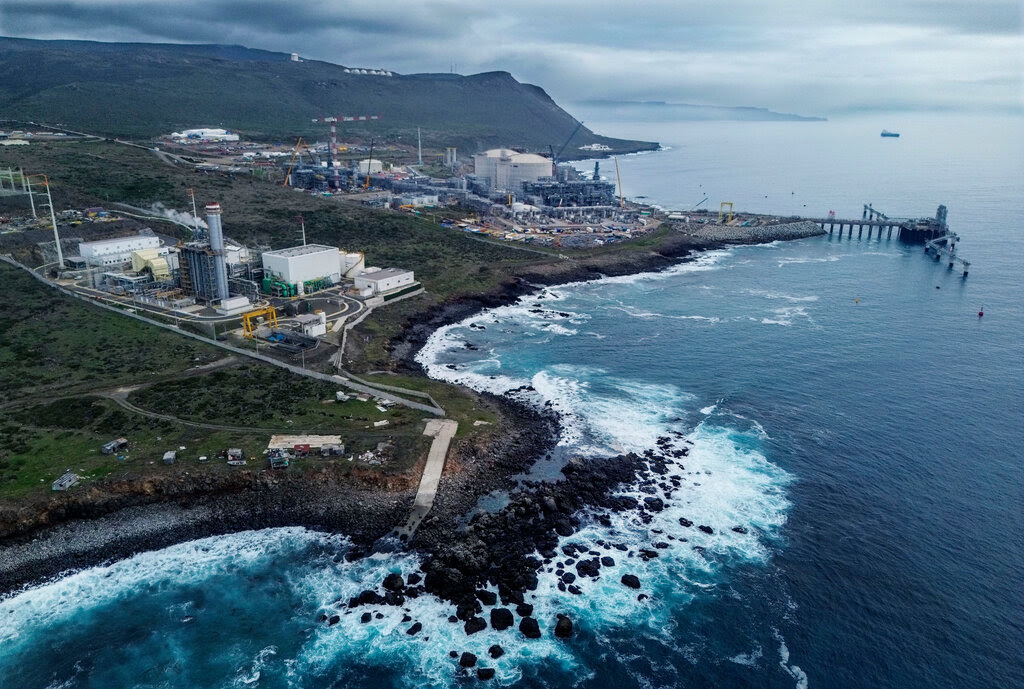Aerial view of a sprawling industrial complex on a rugged coastline with a pier extending over vivid blue ocean.