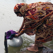 Woman dressed in sari stands on rock at edge of water, dips urn with sari cloth covering opening into water to filter it