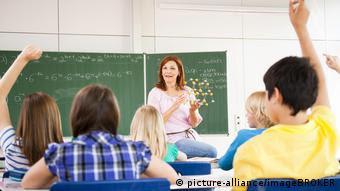 Fotografados de costas, dois alunos aleães levantam as mãos sentados em suas carteiras enquanto professora mostra modelo de ligação química diante de lousa verde na qual estão escritos cálculos com giz