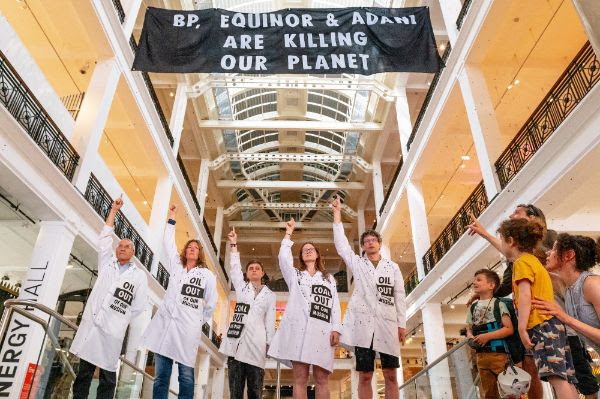 Inside the science museum in London, five people in white lab coats point upwards to a large black banner that says BP, Equinor and Adani are killing our planet