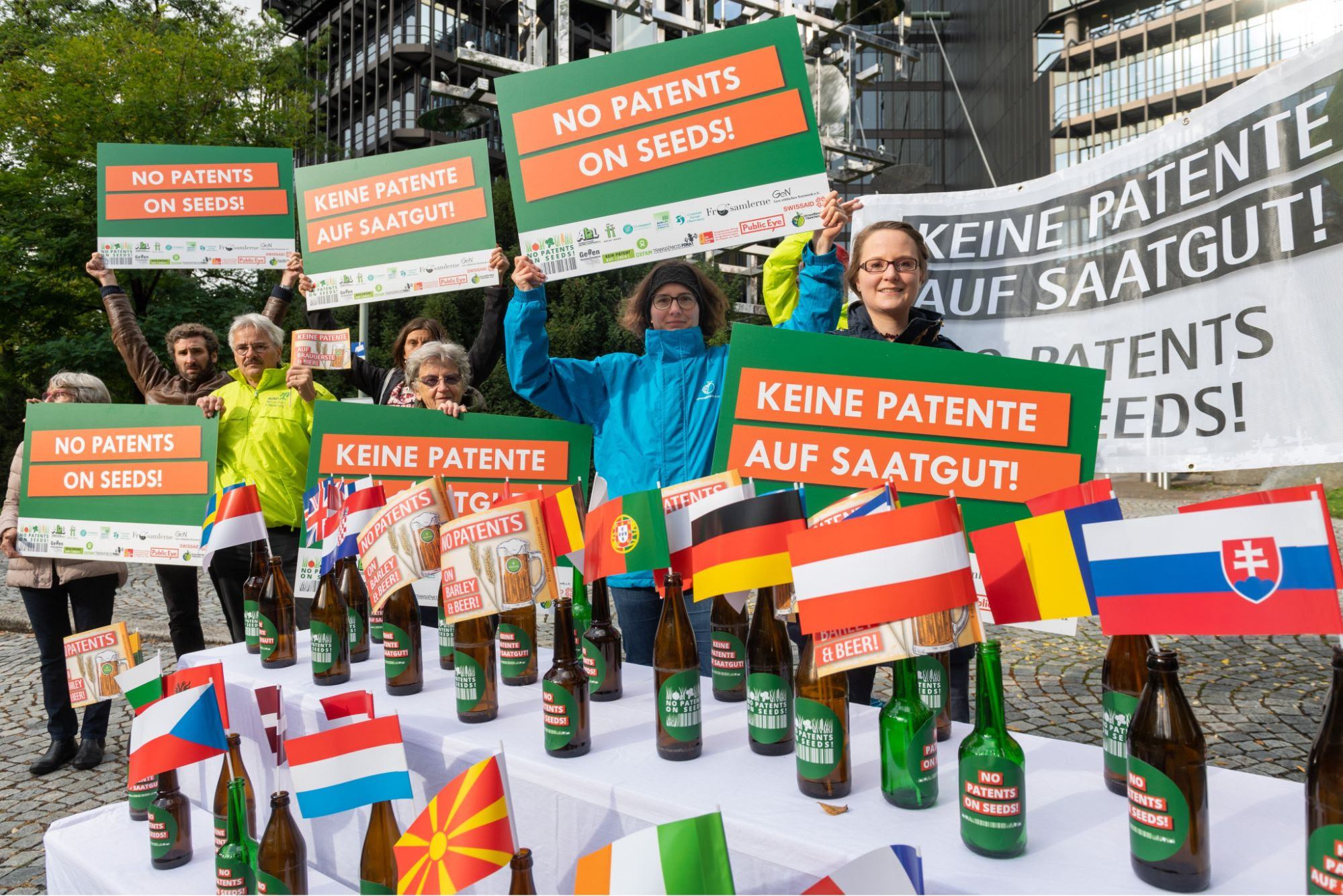 Front of picture, table covered in beer bottles with European flags in them. Behind table to left, group of 6 people holding placards.