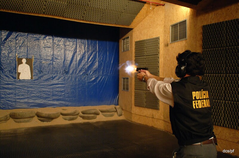 A foto apresenta um agente da Polícia Federal em um stand praticando tiro com uma arma Glock preta; o agente veste um colete preto com "Polícia Federal" escrito em amarelo e fones de proteção, ele está de costas apontando a arma para o alvo fixado em uma parede azul.