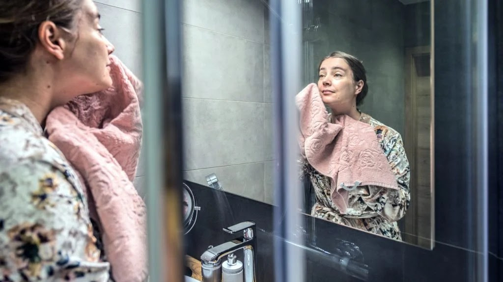 A woman looking in a bathroom mirror as she dries her face with a towel following a treatment for dark spots on the face.