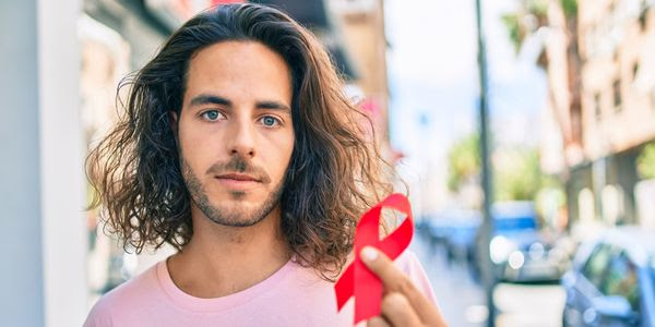 A white man with shoulder-length hair holding up a red ribbon symbolizing HIV/AIDS awareness
