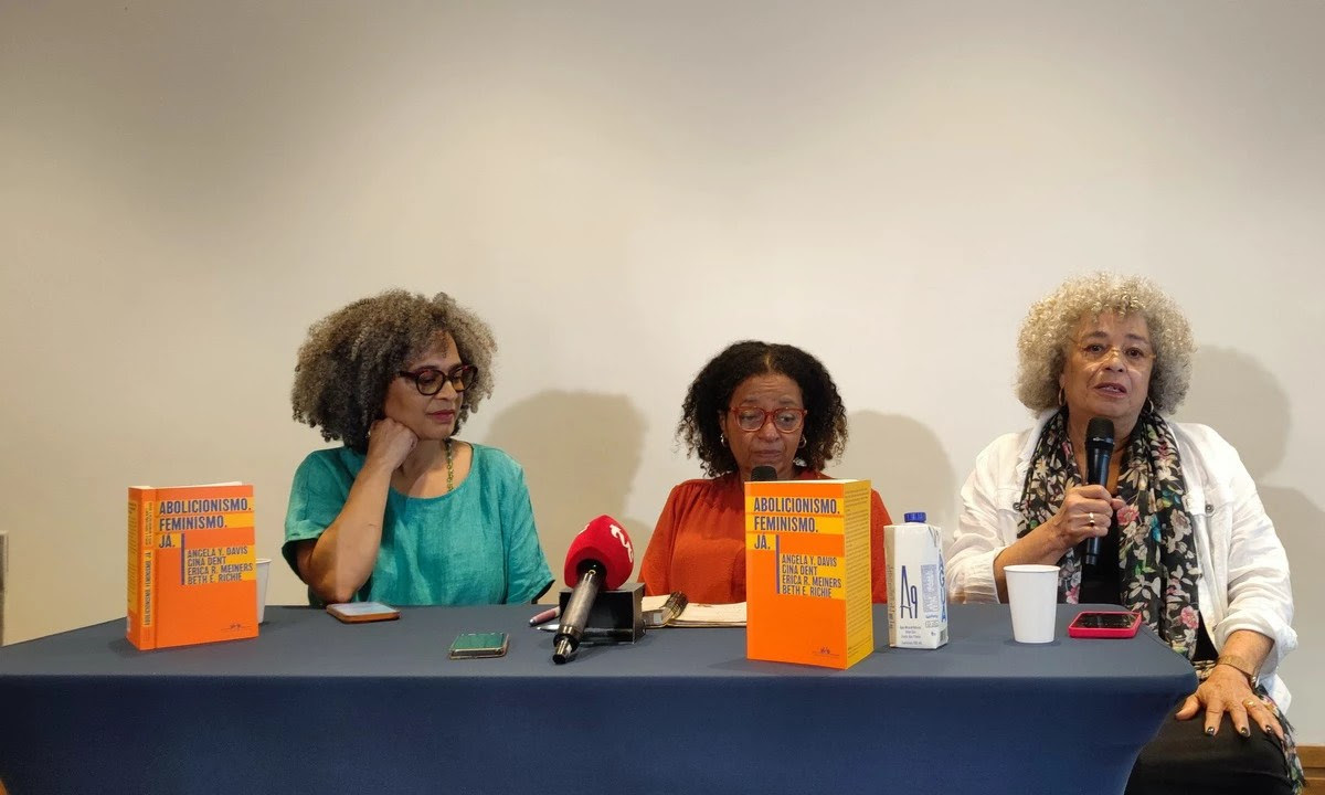 Gina Dent (à esq), Raquel de Souza (ao centro) e Angela Davis (à dir) durante lançamento do livro “Abolicionismo. Feminismo. Já.”. Elas estão sentadas em uma mesa com dois livros de capa laranja expostos