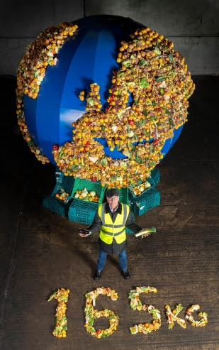Gregg Wallace standing in front of a food waste globe holding a leek and an aubergine. The figure '165kg' is laid out in food waste to represent the amount of food wasted per household per week in the UK