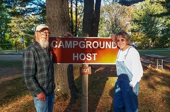 older, smiling man in dark green flannel and baseball cap, and smiling woman in sunglasses and overalls, stand by an orange campground host sign