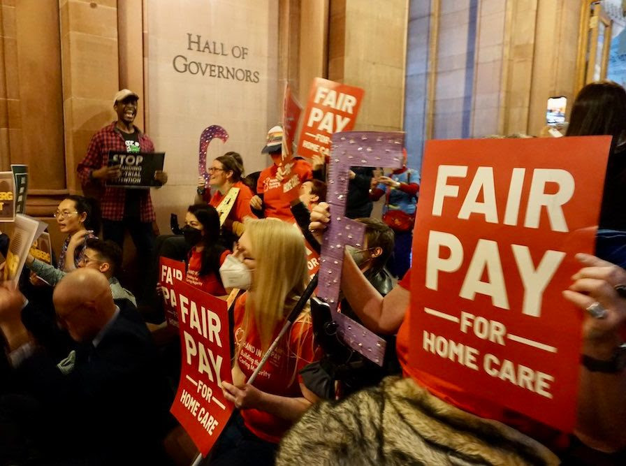 Photo of a protest inside the Hall of Governors. People are holding fair pay for home care signs