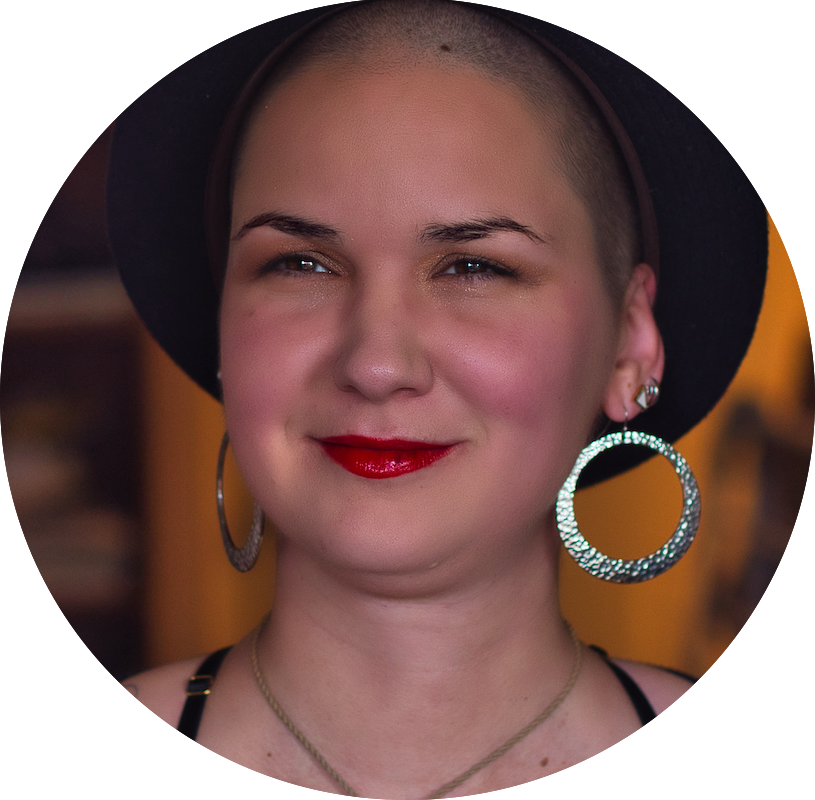 Karina, a woman with shaved hair is wearing a black hat and large silver earrings.