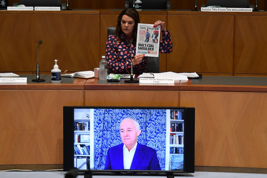 A dark-haired woman holds up a newspaper at a hearing room above a monitor with an older man in blue suit.