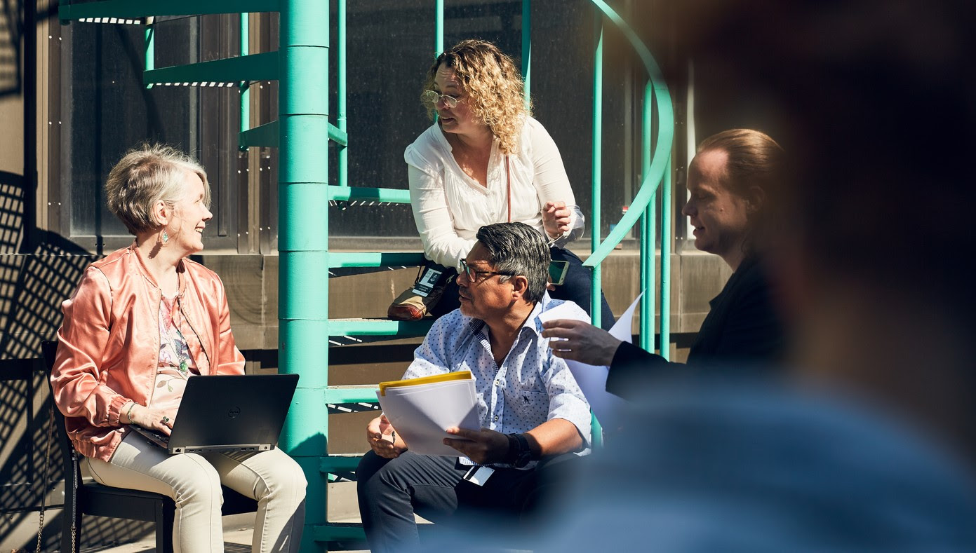 Fyra personer med datorer och papper är samlade vid en spiraltrappa och verkar inbegripna i ett samtal.