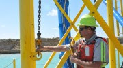 A worker in a hard hat stands on a metal structure, holding a chain.