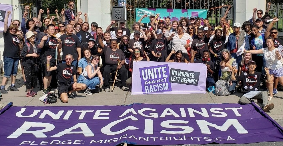 Close to 80 people wearing $15 & Fairness shirts pose behind
a banner that reads Unite Against Racism