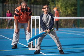 A young visually impaired boy playing adapted cricket with a coach behind him watching