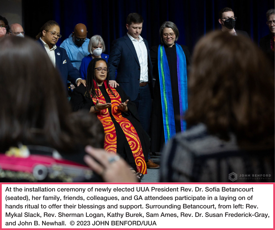 Rev. Sofia Betancourt is seated wearing a red and gold colored stole, with people standing in a wide circle around her