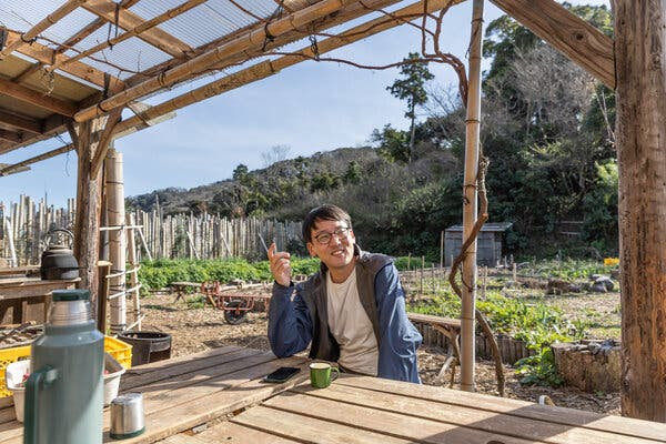 This color photo shows a youthful-looking man in glasses and wearing a blue jacket over a white T-shirt sitting at an outdoor wooden table beneath a wooden pergola, his cellphone and a green coffee mug before him. Behind him we can see rows of plants and some garden equipment.