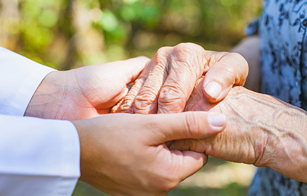 A close-up of a nurse’s hands holding an elderly woman’s trembling hands.