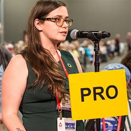 a delegate for the 2019 General Assembly speaking at a microphone with a sign attached that says PRO