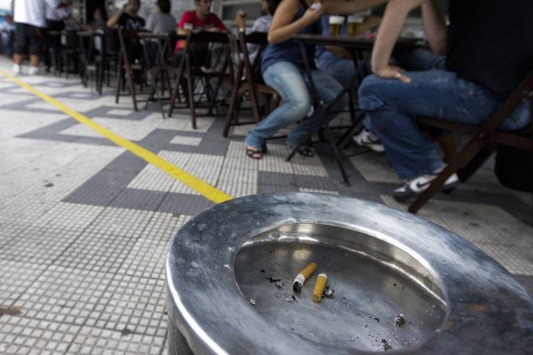 Cinzeiros na calçada da Vila Madalena, em São Paulo (SP). Ao fundo, pessoas sentadas em um bar