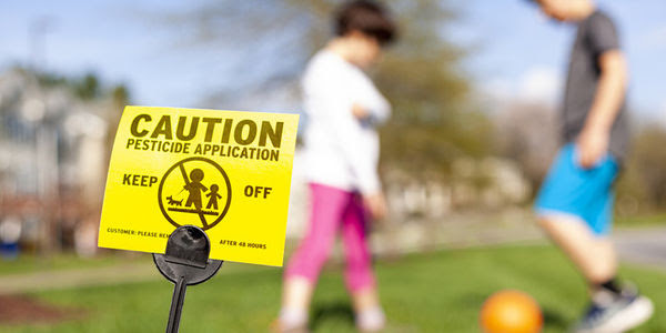 Two children play soccer on a yard with a ''Caution: Pesticide Application'' sign.