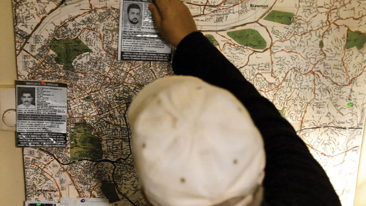 [(테)에러 스틸컷] 지도 위에 두 장의 얼굴 사진이 붙어 있다. 모자를 쓴 사람이 한 장의 사진에 손을 대고 있다.