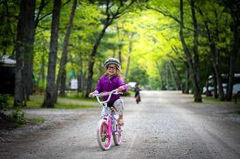 Bundled-up little girl, smiling, riding her bike down a gravel trail, bright green trees in background