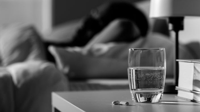 Black and white image of a glass of water and medication on a night stand. You can see a person in the background laying down but it is extremely out of focus.