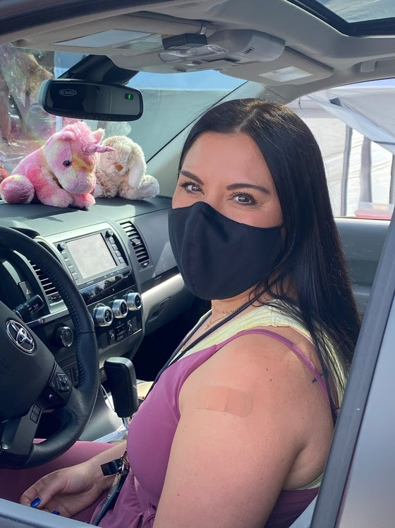 Woman sits in car with bandage on shoulder wearing a black face mask. The dashboard has a stuffed rainbow unicorn and a stuffed bunny toy.
