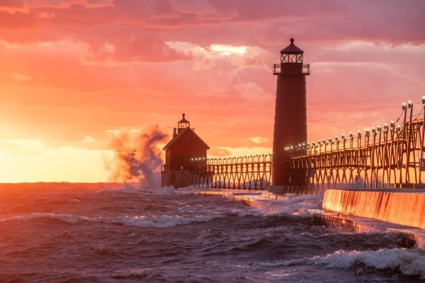 A massive wave sprays up against the side of a lighthouse in the red haze of a summer sunset.