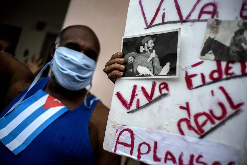 Homem com máscara de proteção comemora o Dia Internacional do Trabalho enquanto outra pessoa segura uma placa com fotos de heróis revolucionários e a mensagem 'Viva Fidel e Raul', em Havana, Cuba — Foto: Ramon Espinosa/AP