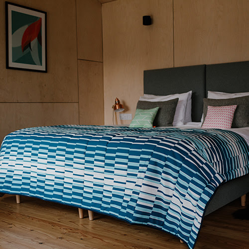 A photograph of a bed in a wooden cabin with a large blue quilt by Laura Spring.