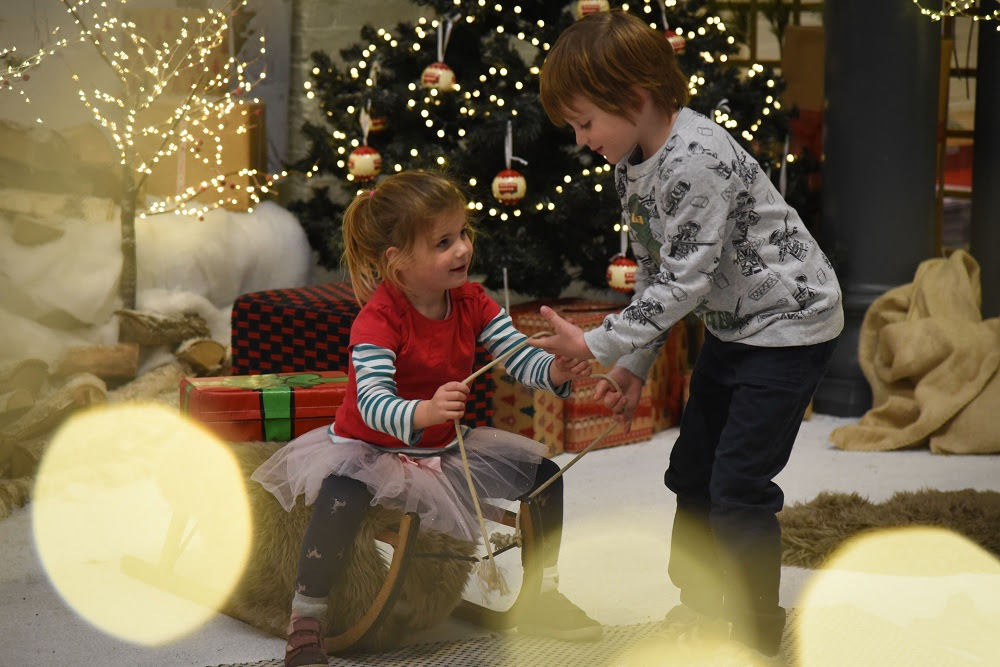 Two children playing on a wooden sledge in front of a Christmas tree