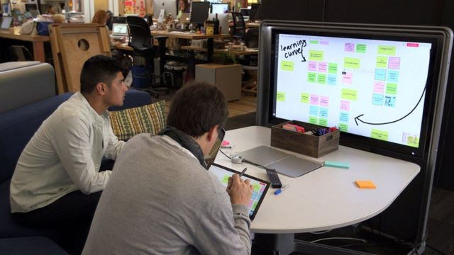 Em escritório, dois homens em frente a tela com lousa virtual