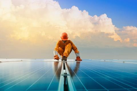 A person in orange overalls and hard hat kneels on top of rooftop solar panels.