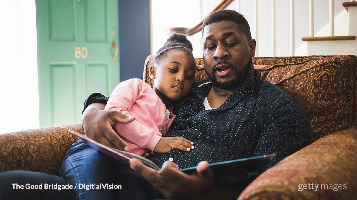 A father reads a picture book aloud as his daughter sits on his lap and looks at the picture; Father and daughter are both Black.