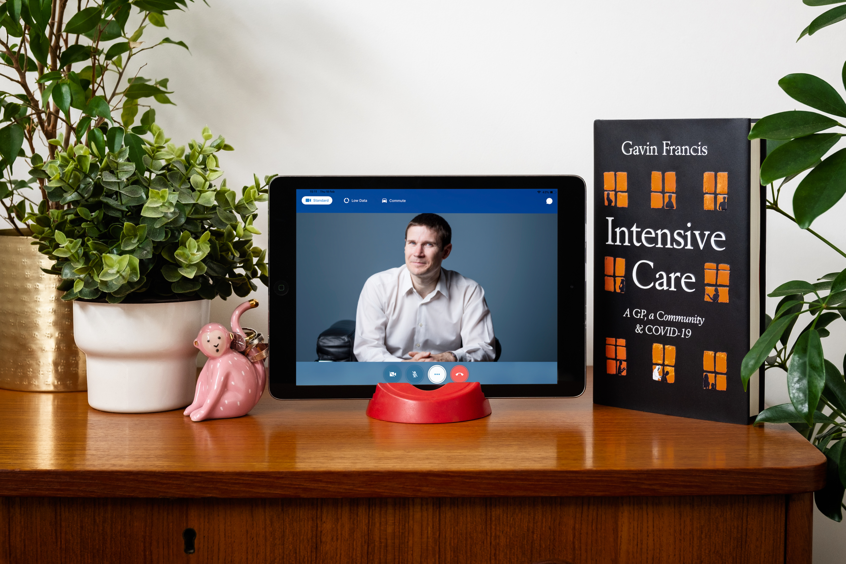 An image of tablet computer which displays Dr Gavin Francis seated in a chair. His Intensive Care book is stood upright next to the tablet surrounded by a pink toy monkey and few plants.