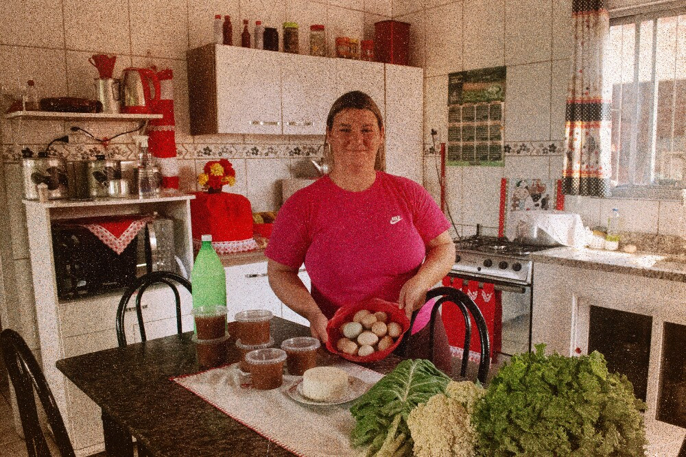 Uma mulher branca está em uma cozinha, ela segura um pote com ovos; na mesa à sua frente estão algumas verduras que ela colheu de sua horta