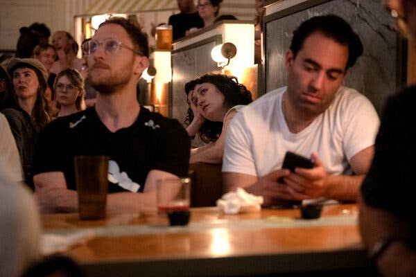 People in a restaurant watch the debate on a television that is out of view. A man at the table in front is looking at his phone. 
