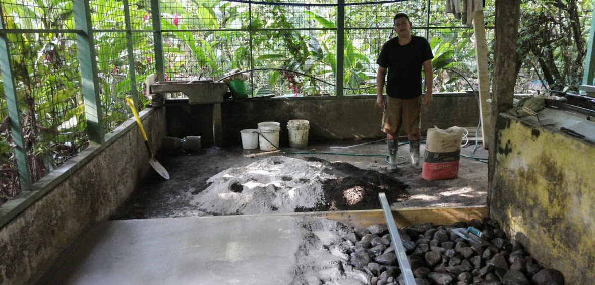 A worker stands behind a half-finished concrete floor with jungle in the background