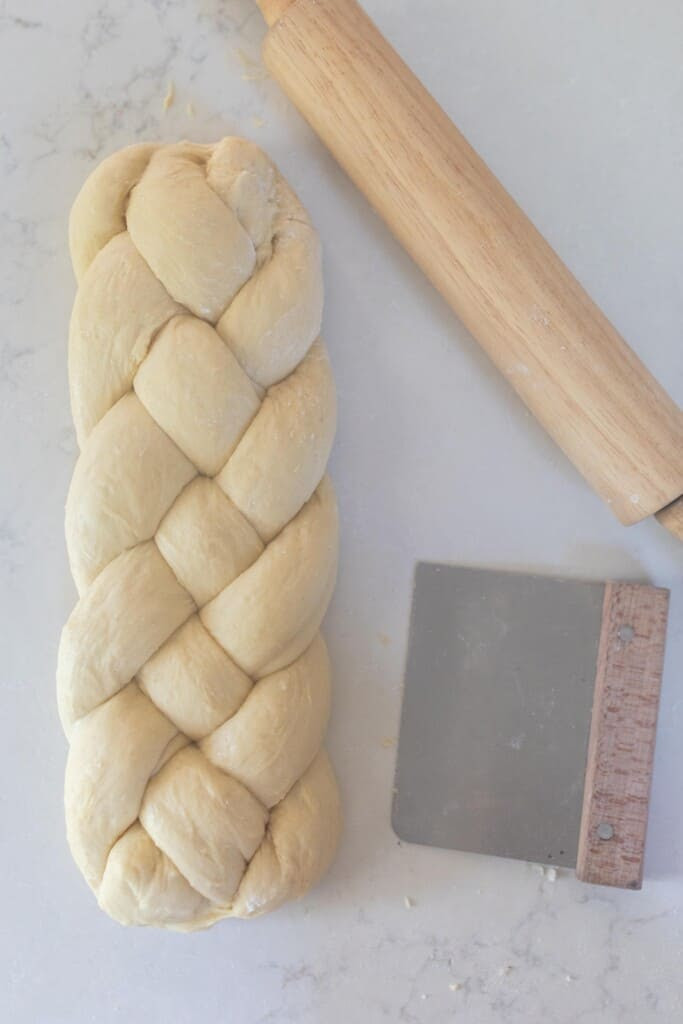 a loaf of braiding sourdough bread dough on a white countertop with a bench scraper and rolling pin to the right