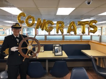 Photo of woman captain holding a ship's wheel in front of balloons that spell out "Congrats"