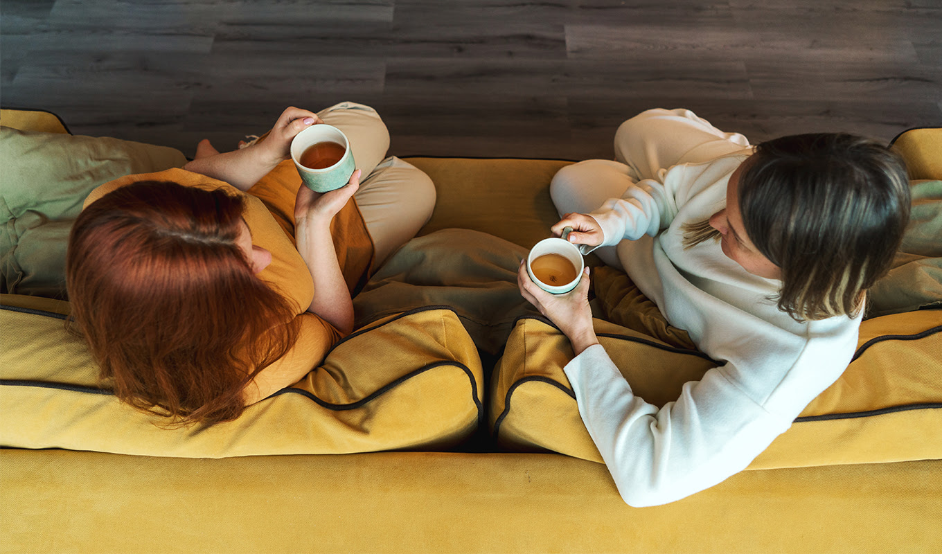 green tea vs. black tea Two women are sitting and talking on a yellow couch with teas in their hands