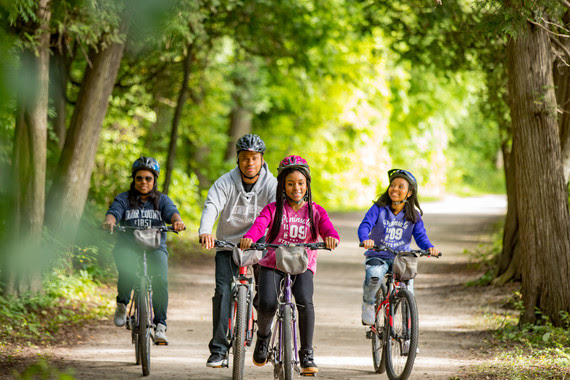 A family of four smiles as they ride bikes towards the camera on a trail. 