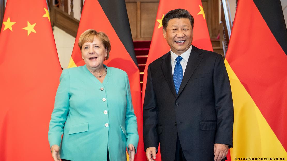 Foto registra Angela Merkel e Xi Jinping, ambos sorrindo, em frente a bandeiras da Alemanha e da China.