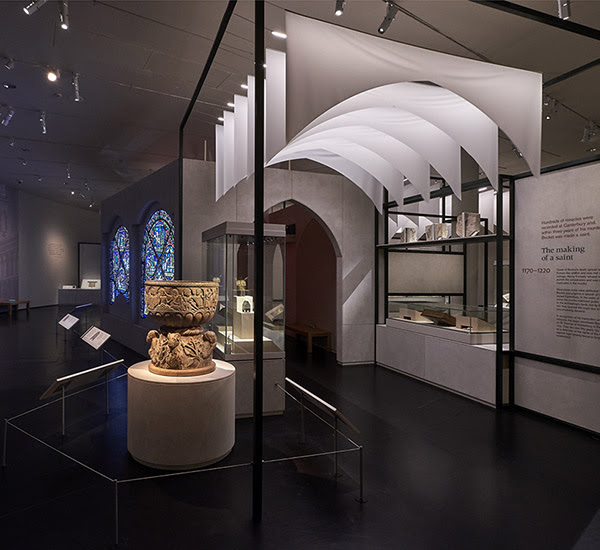 Inside 'Thomas Becket: murder and the making of a saint' showing stained glass, a stone font, and other objects on display. Hanging drapes form the shape of an arched corridor.