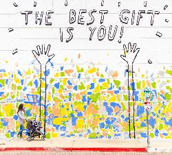 Fence decorated with many colors, almost like flowers or confetti, with arms drawn reaching upward toward text "The best gift is you." Photo by Dakota Corbin via Unsplash.