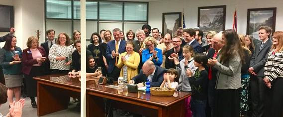 A large crowd is gathered behind the governor to watch him sign the Computer Science Education bill into law in a meeting room at the Jonah Financial Center. Observers include State Superintendent Jillian Balow, state legislators, tech company representatives, Array School students, and other supporters of the bill.