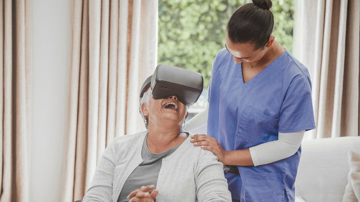 An older woman sitting down in a chair wearing a VR headset. She is smiling big while the nurse stands behind her and helps her.